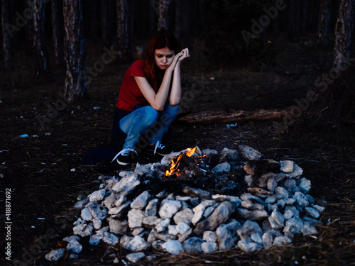 A romantic woman in jeans and a T-shirt sits near a campfire in nature