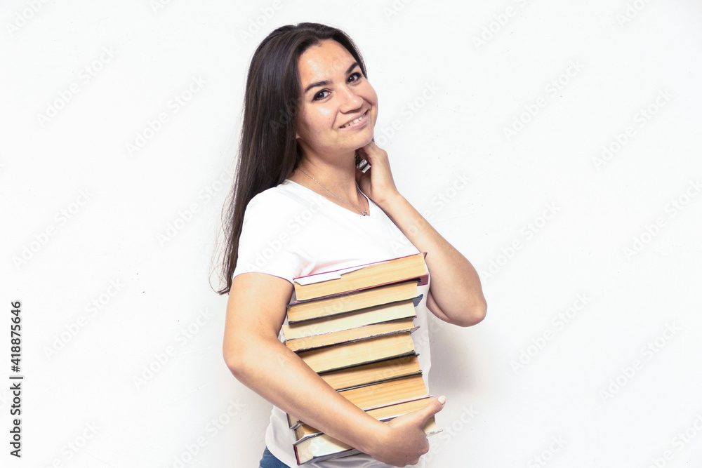 Young smiling student woman in white t-shirt holding textbooks and books in a pile on a white backdrop. College education concept. Positive emotions, facial expression Young smiling student woman in white t-shirt holding textbooks and books in a pile on a white backdrop. College education concept. Positive emotions, facial expression