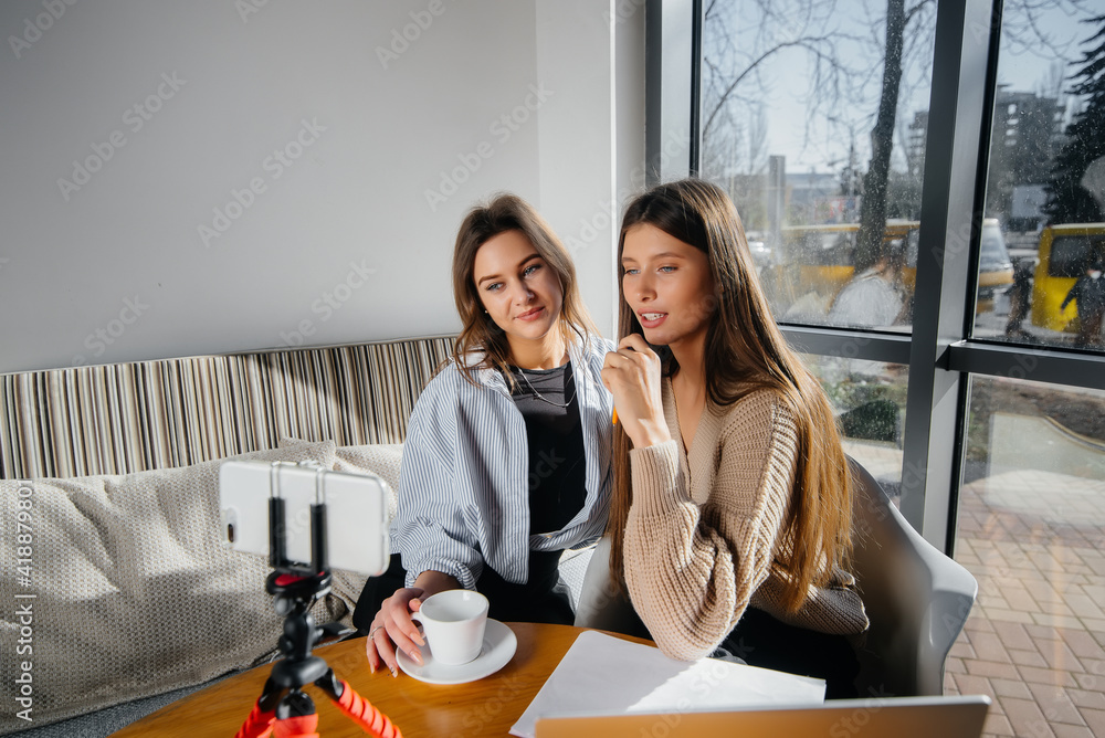 Fototapeta premium Two young beautiful girls are sitting in a cafe, recording video blogs and communicating on social networks.