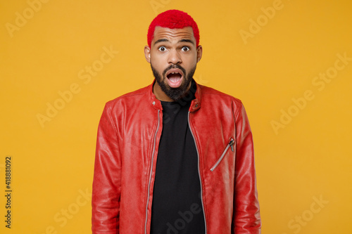 Young african dark-skinned shocked indignant surprised man 20s with funky trendy pink hairdo wear red leather jacket look camera with opened mouth isolated on yellow orange background studio portrait.