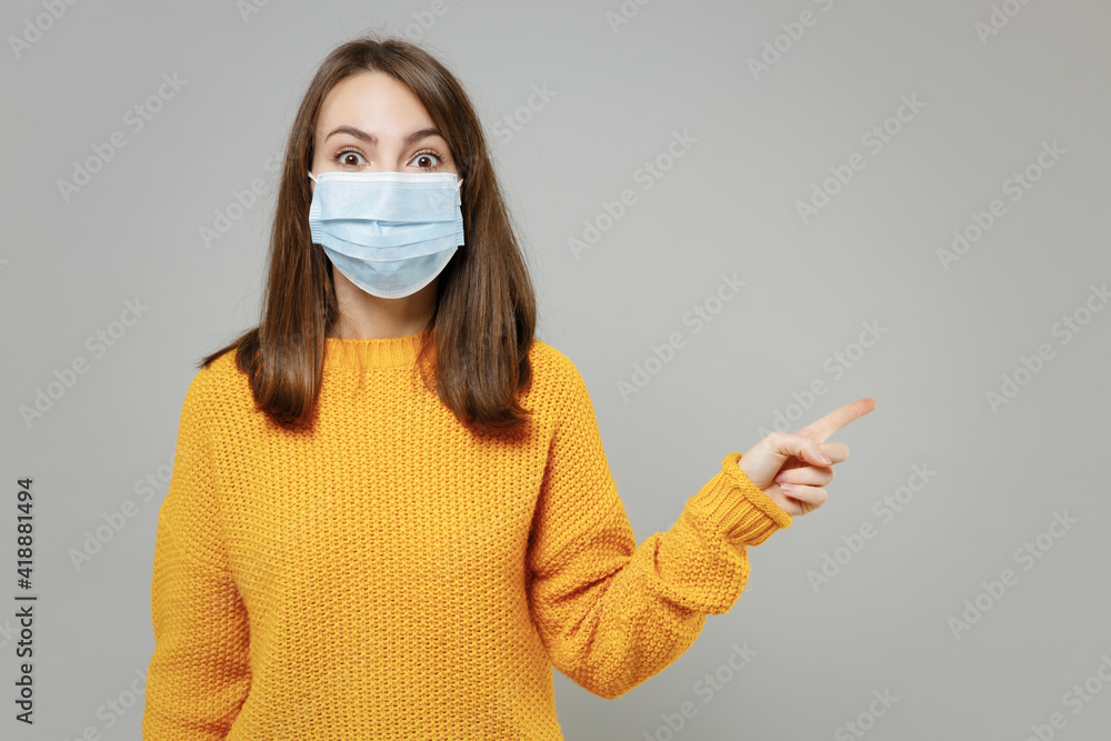 Young woman in yellow sweater in sterile face mask to safe from coronavirus virus covid-19 during pandemic quarantine point index finger aside on workspace isolated on grey background studio portrait Young woman in yellow sweater in sterile face mask to safe from coronavirus virus covid-19 during pandemic quarantine point index finger aside on workspace isolated on grey background studio portrait