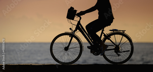Silhouette of a woman traveler with her bicycle rides on sea coastline, sunset sky in sunny summer day on beach