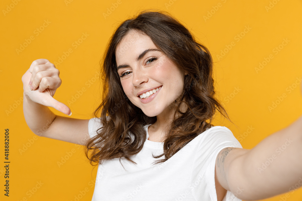 Close up young confident lucky smiling friendly caucasian woman in white casual tshirt do selfie shot on mobile phone point thumb finger on herself isolated on yellow color background studio portrait Close up young confident lucky smiling friendly caucasian woman in white casual tshirt do selfie shot on mobile phone point thumb finger on herself isolated on yellow color background studio portrait