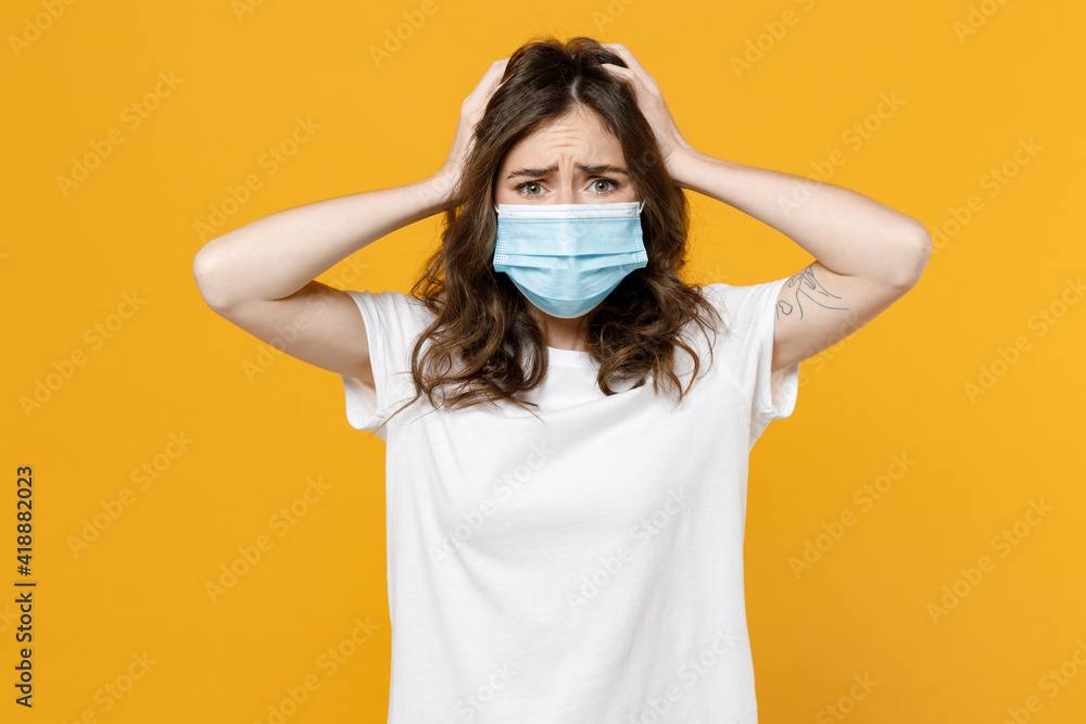 Young troubled sad disappointed woman in white basic t-shirt in sterile face mask from coronavirus virus covid-19 during pandemic quarantine hold head isolated on yellow orange color background. Young troubled sad disappointed woman in white basic t-shirt in sterile face mask from coronavirus virus covid-19 during pandemic quarantine hold head isolated on yellow orange color background.