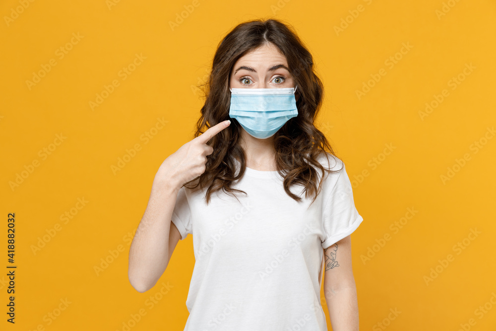 Young woman 20s wearing white basic blank print design t-shirt point index finger on sterile face mask to safe from coronavirus virus covid-19 pandemic quarantine isolated on yellow background studio. Young woman 20s wearing white basic blank print design t-shirt point index finger on sterile face mask to safe from coronavirus virus covid-19 pandemic quarantine isolated on yellow background studio.