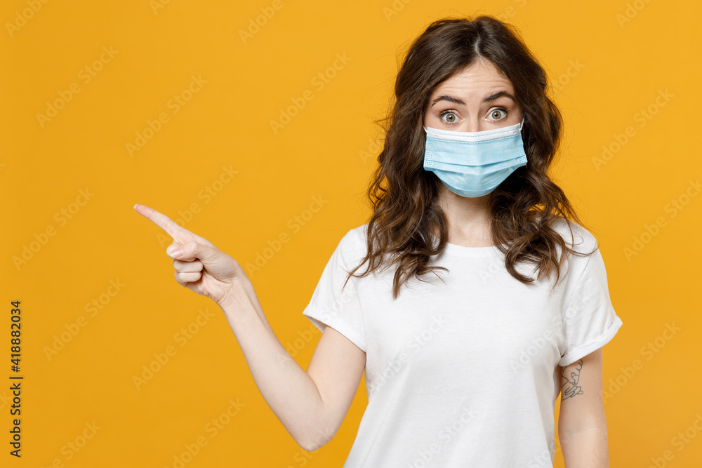 Young woman in white basic blank print design t-shirt in sterile face mask from coronavirus virus covid19 pandemic quarantine point finger aside on workspace area isolated on yellow orange background. Young woman in white basic blank print design t-shirt in sterile face mask from coronavirus virus covid19 pandemic quarantine point finger aside on workspace area isolated on yellow orange background.