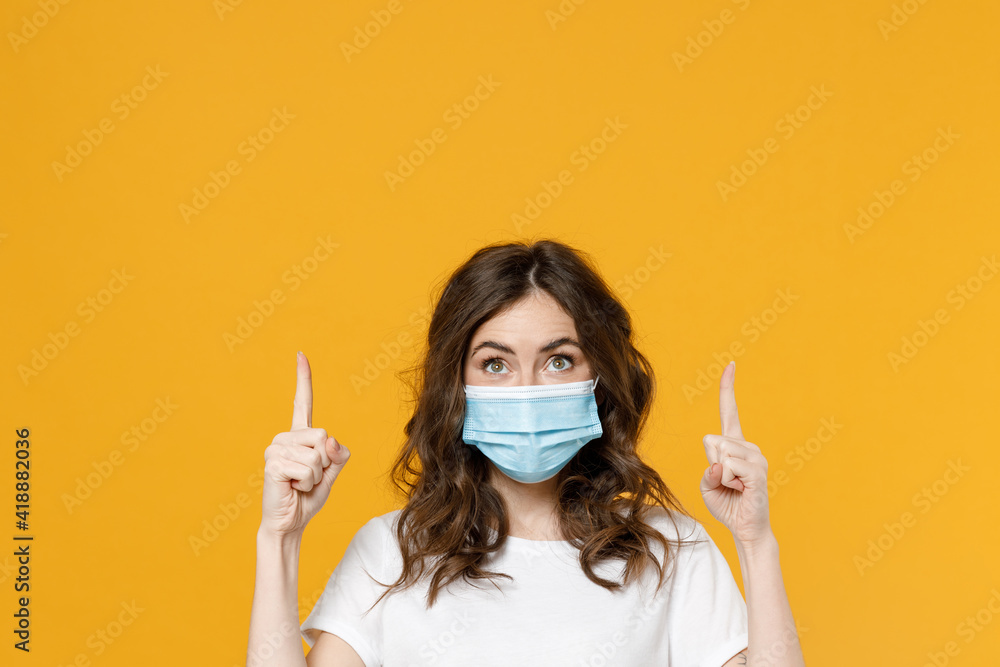 Young woman in white basic blank print design t-shirt in sterile face mask from coronavirus virus covid-19 pandemic quarantine point finger up on workspace area isolated on yellow orange background. Young woman in white basic blank print design t-shirt in sterile face mask from coronavirus virus covid-19 pandemic quarantine point finger up on workspace area isolated on yellow orange background.