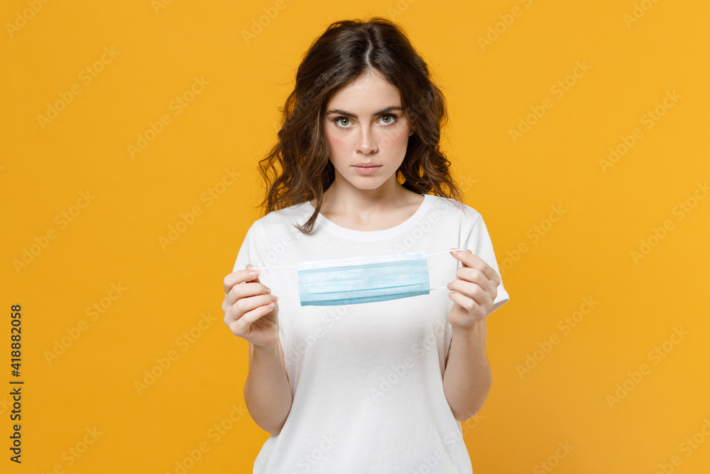 Young woman in white basic blank print design t-shirt hold sterile face mask safe from coronavirus virus covid-19 during pandemic quarantine look camera isolated on yellow background studio portrait. Young woman in white basic blank print design t-shirt hold sterile face mask safe from coronavirus virus covid-19 during pandemic quarantine look camera isolated on yellow background studio portrait.