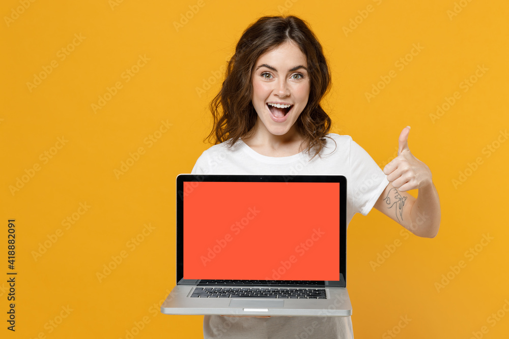 Naklejka premium Young fun freelancer copywriter student woman in white basic t-shirt hold laptop pc computer with blank screen workspace area show thumb up like gesture isolated on yellow background studio portrait.