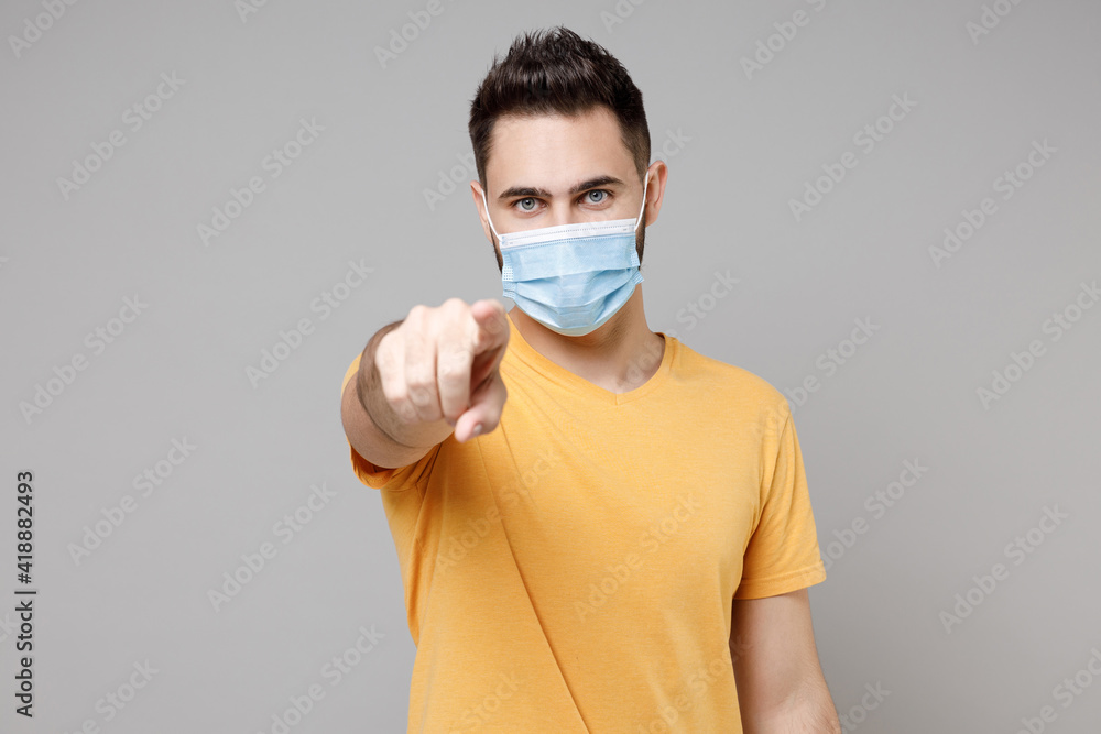 Young caucasian man 20s in yellow t-shirt wear sterile face mask to safe coronavirus virus covid-19, pandemic quarantine point index finger camera on you isolated on grey background studio portrait. Young caucasian man 20s in yellow t-shirt wear sterile face mask to safe coronavirus virus covid-19, pandemic quarantine point index finger camera on you isolated on grey background studio portrait.