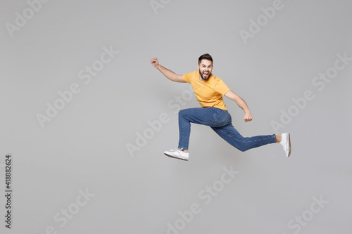 Full length of young bearded sport active student hurrying man 20s wearing yellow basic t-shirt jump high run away fast looking camera outstretched hands isolated on grey background studio portrait.