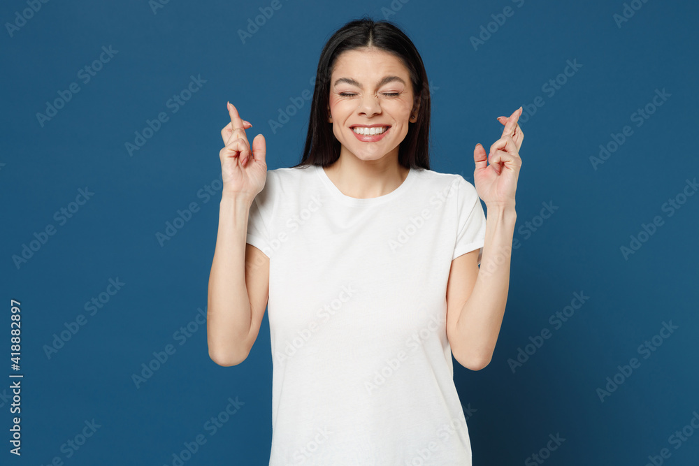 Young dreamful latin happy woman 20s wearing white casual basic t-shirt wait for special moment keeping fingers crossed making wish eyes closed isolated on dark blue color background studio portrait. Young dreamful latin happy woman 20s wearing white casual basic t-shirt wait for special moment keeping fingers crossed making wish eyes closed isolated on dark blue color background studio portrait.