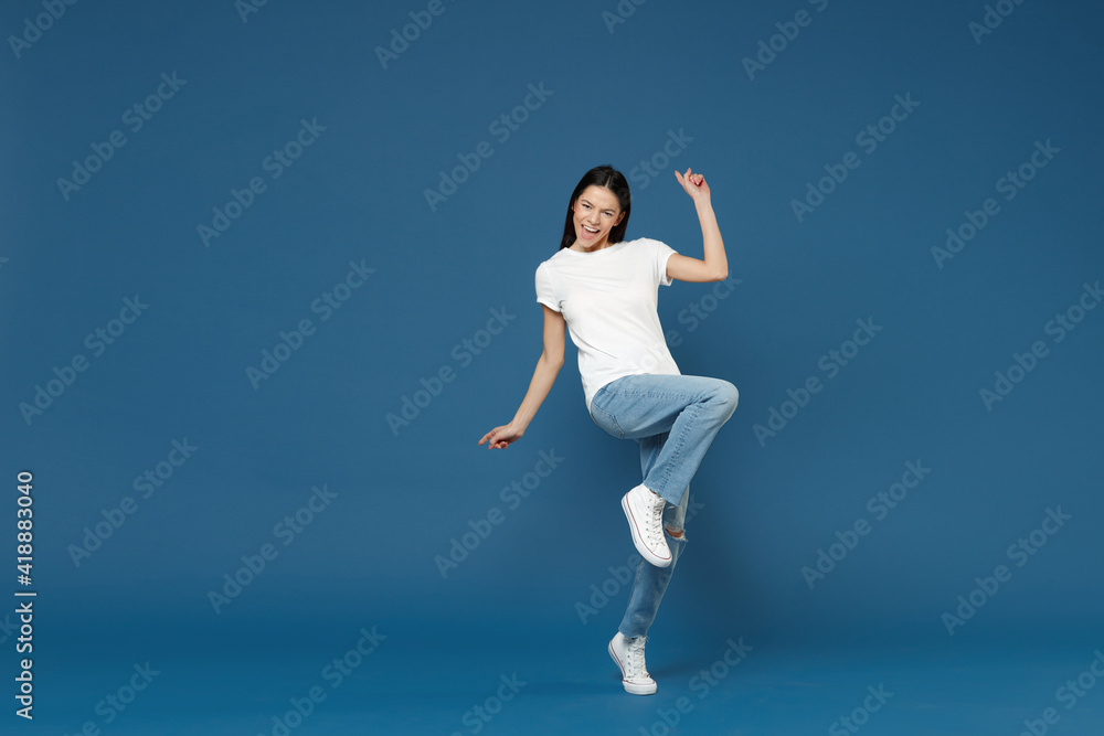 Full length young relaxed overjoyed brunette latin woman 20s wearing white casual basic t-shirt leaning over with raised up leg looking camera isolated on dark blue color background studio portrait.