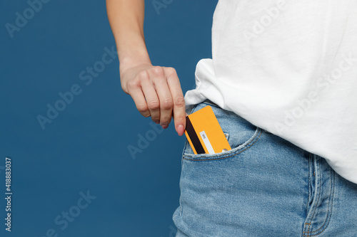 Close up cropped photo portrait shot of caucasian female hand arm putting credit bank card in jeans pants denim pocket isolated on dark blue background studio portrait. Money finance currency concept. photo