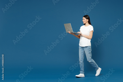 Full length young happy brunette freelancer latin woman 20s in white basic t-shirt hold laptop pc computer chat typing on keyboard use social internet isolated on dark blue background studio portrait.
