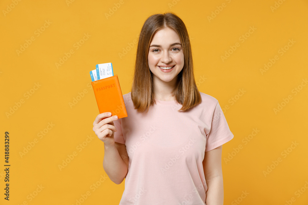 Traveler tourist woman 20s in basic pastel pink t-shirt hold passport with tickets isolated on yellow orange background. Passenger traveling abroad on weekends getaway. Air flight journey concept. Traveler tourist woman 20s in basic pastel pink t-shirt hold passport with tickets isolated on yellow orange background. Passenger traveling abroad on weekends getaway. Air flight journey concept.