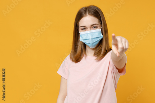 Young woman in basic pastel pink t-shirt, blank print design, sterile face mask to safe from coronavirus covid-19 pandemic quarantine point index finger camera on you isolated on yellow background