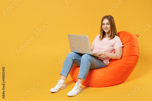 Full length of young student woman 20s in basic pastel pink t-shirt, jeans sitting in orange bean bag chair holding laptop pc computer surfing internet in lounge zone isolated on yellow background.
