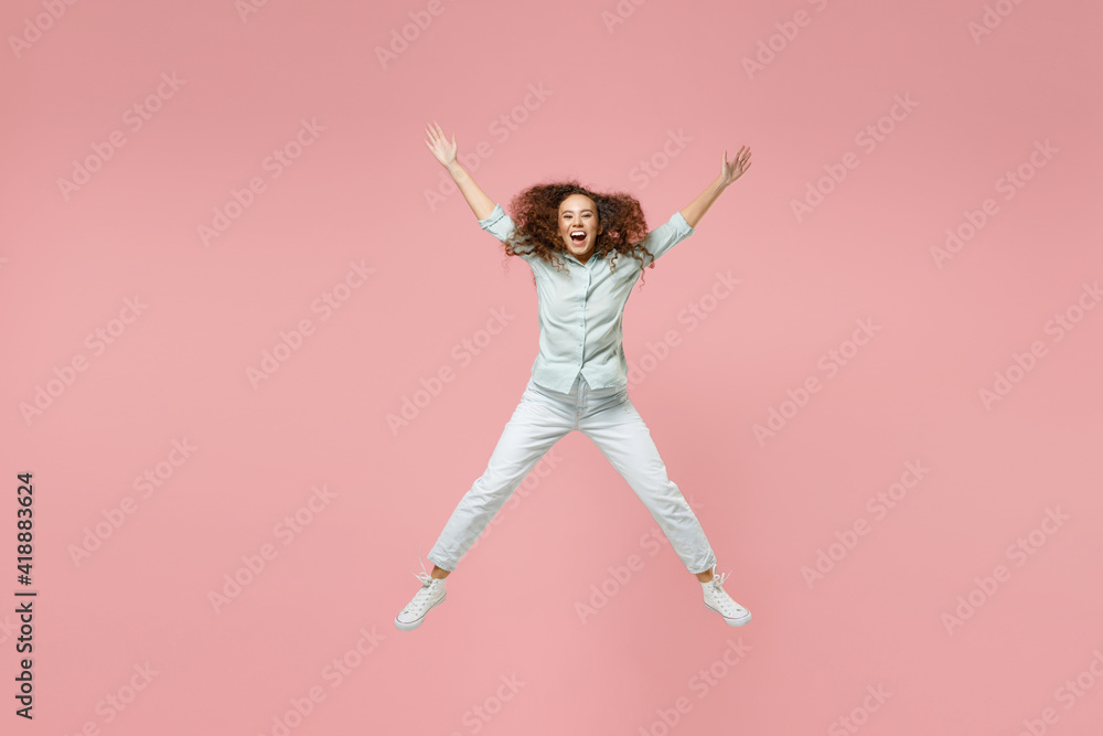 Full length young black african fun happy surprised overjoyed excited joyful woman 20s in blue shirt jumping high with outstretched hands legs isolated on pastel pink color background studio portrait.