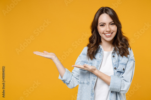 Young smiling woman promoter in denim shirt white t-shirt recommend suggest select advert point index finger aside on workspace commercial promo area mock up copy space isolated on yellow background.