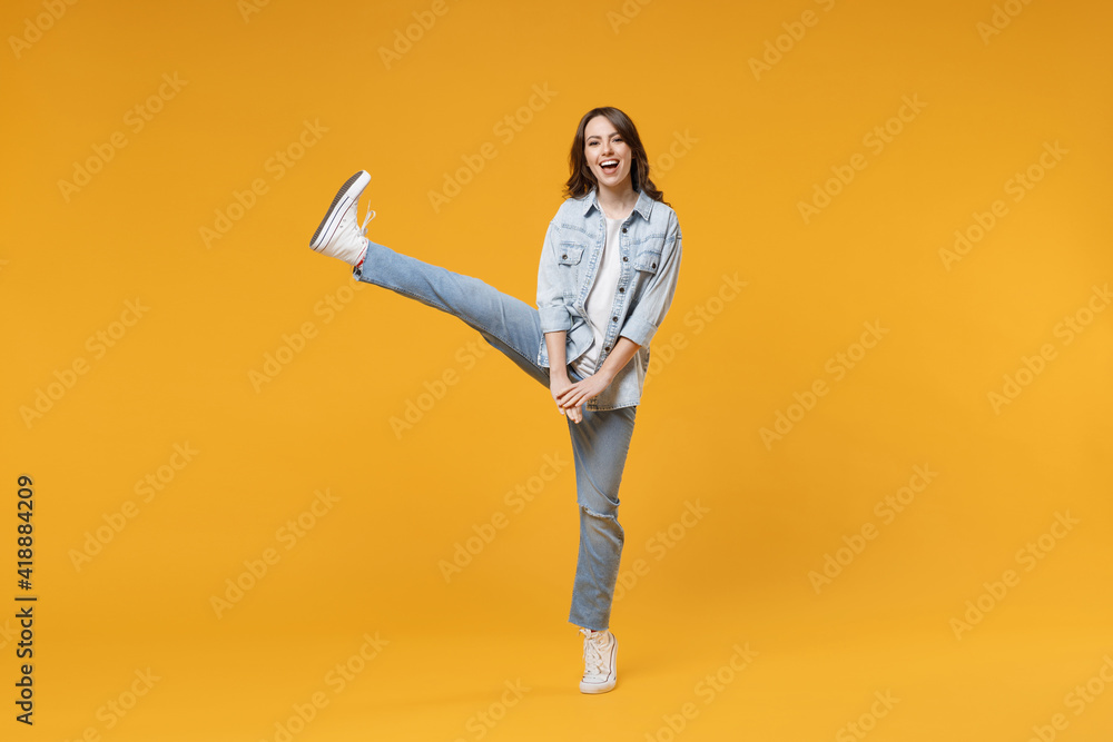Full length of young caucasian relaxed fun student happy stylish woman 20s in denim shirt white t-shirt stand with outstretched raised up leg look camera isolated on yellow background studio portrait Full length of young caucasian relaxed fun student happy stylish woman 20s in denim shirt white t-shirt stand with outstretched raised up leg look camera isolated on yellow background studio portrait