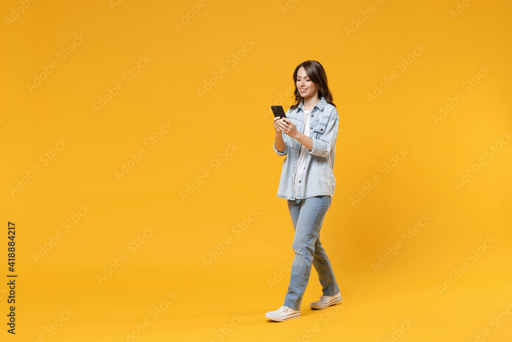 Full length of young smiling friendly fun student happy woman 20s wear casual denim shirt white t-shirt holding mobile cell phone chatting walking going isolated on yellow background studio portrait. - obrazy, fototapety, plakaty 