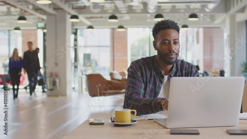 Dolly shot of casually dressed businessman working on laptop taking coffee break at desk in modern open plan start up office with colleagues in background - shot in slow motion