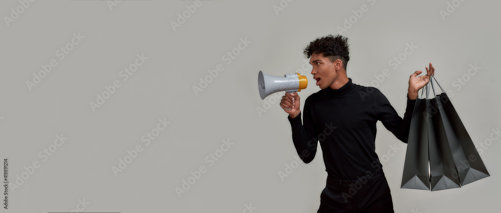 Obraz premium Stylish young guy dressed elegantly in black screaming in megaphone, making sale announcement, posing with shopping bags isolated over gray background
