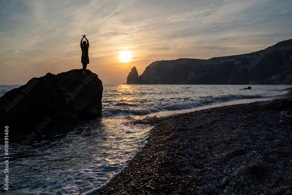 A girl on the beach watches the sunset on the sea. She stands on a rock in the sea and holds her hands up. The sun is setting behind the mountains.