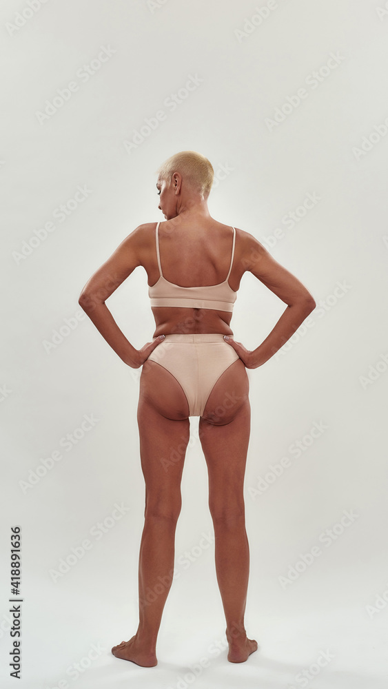 Rear view of a mature woman in underwear keeping arms on heeps while posing  half naked against light background, full length Photos | Adobe Stock