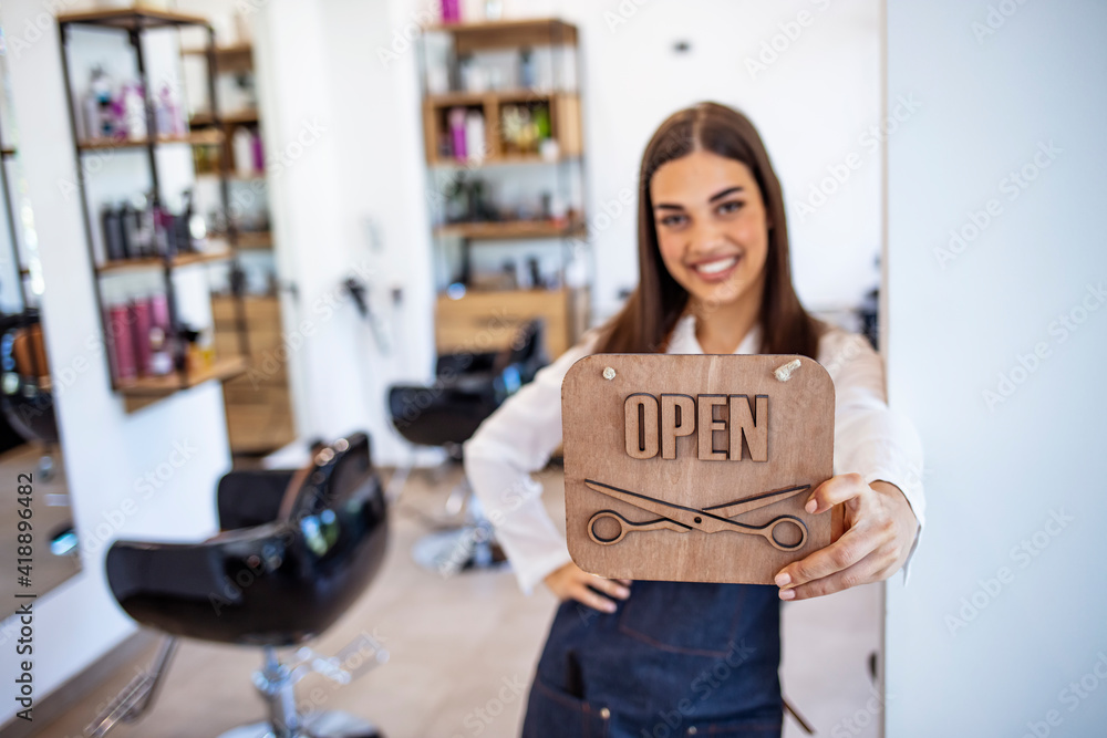 Smiling brunette owner of salon standing with sign open. Smiling owner of hair salon standing with sign open and leaning on glass door. Young female business owner standing outside her salon shop