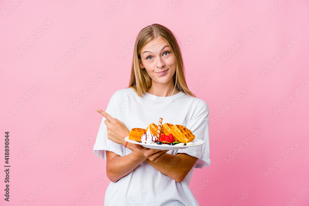 Young russian woman eating a waffle isolated points sideways, is trying to choose between two options. Young russian woman eating a waffle isolated points sideways, is trying to choose between two options.
