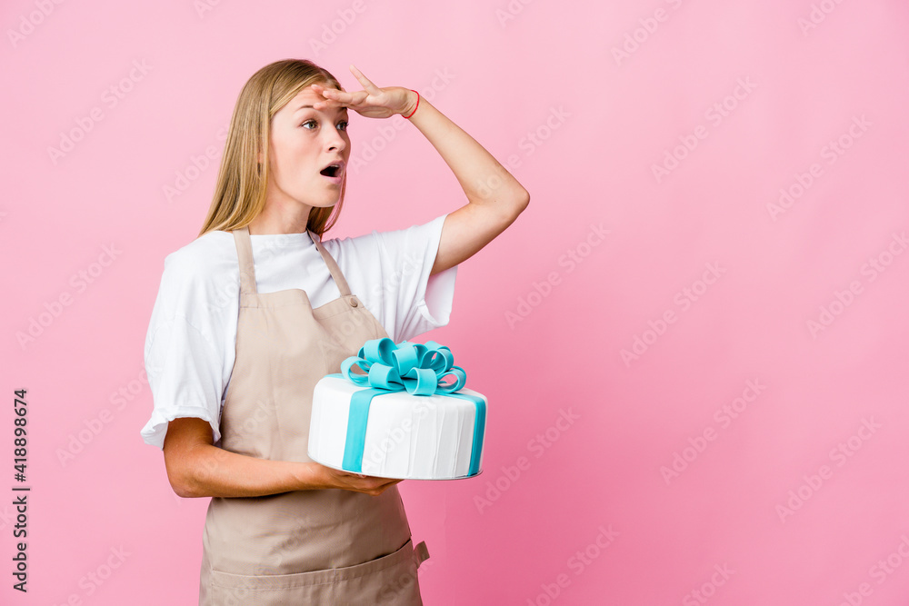 Young russian baker woman holding a delicious cake looking far away keeping hand on forehead. Young russian baker woman holding a delicious cake looking far away keeping hand on forehead.