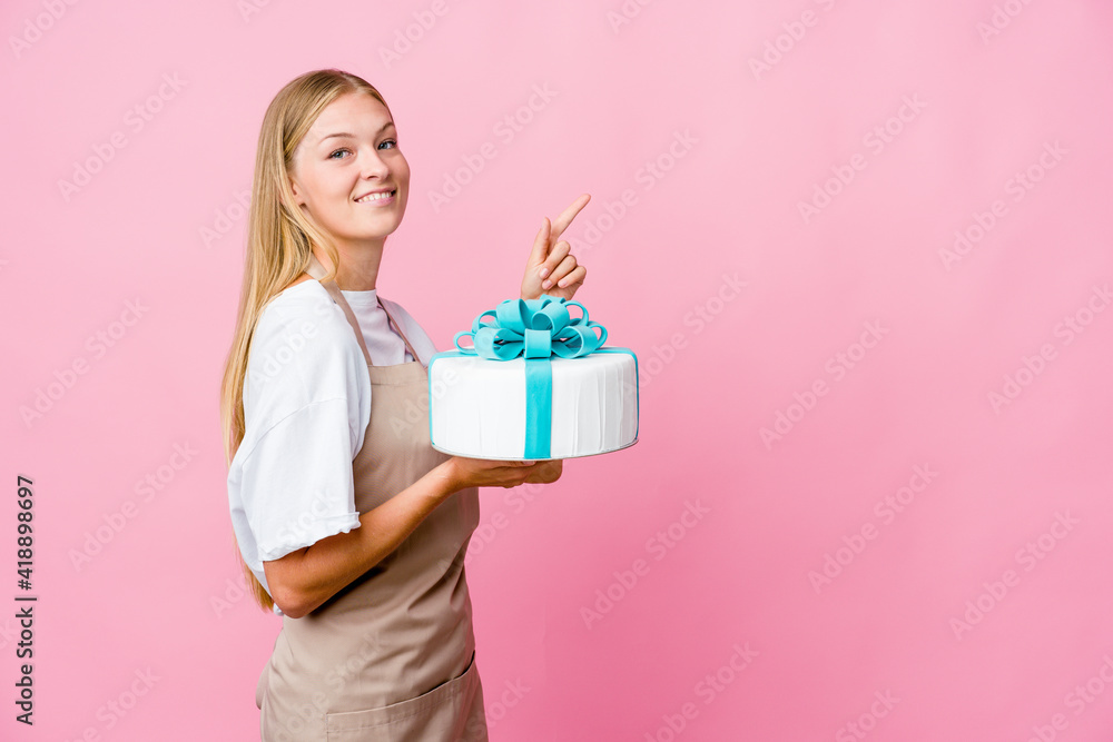Young russian baker woman holding a delicious cake excited pointing with forefingers away. Young russian baker woman holding a delicious cake excited pointing with forefingers away.