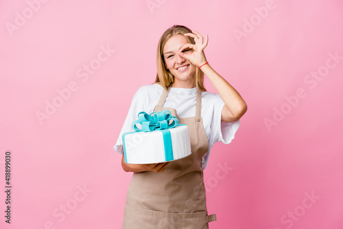 Young russian baker woman holding a delicious cake excited keeping ok gesture on eye.