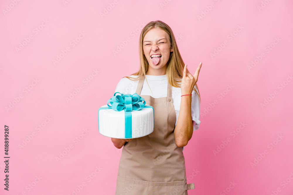 Young russian baker woman holding a delicious cake showing rock gesture with fingers Young russian baker woman holding a delicious cake showing rock gesture with fingers