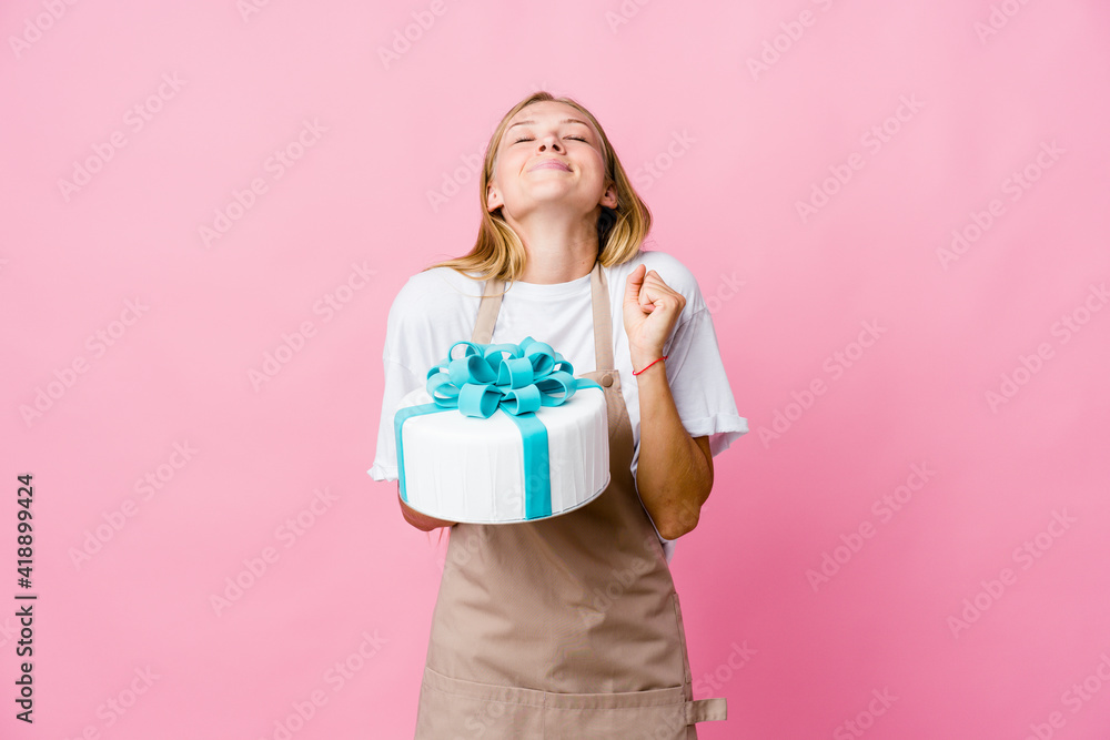 Young russian baker woman holding a delicious cake raising fist, feeling happy and successful. Victory concept. Young russian baker woman holding a delicious cake raising fist, feeling happy and successful. Victory concept.