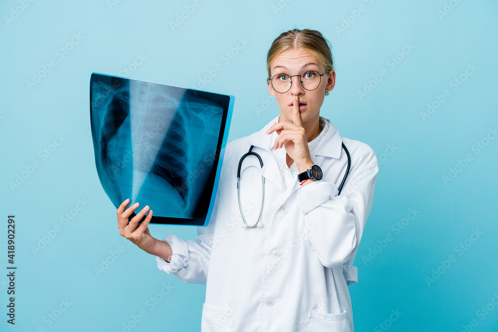 Young russian doctor woman holding a bone scan on blue keeping a secret or asking for silence. Young russian doctor woman holding a bone scan on blue keeping a secret or asking for silence.