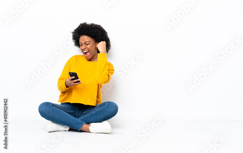 Young African American woman sitting on the floor with phone in victory position © luismolinero
