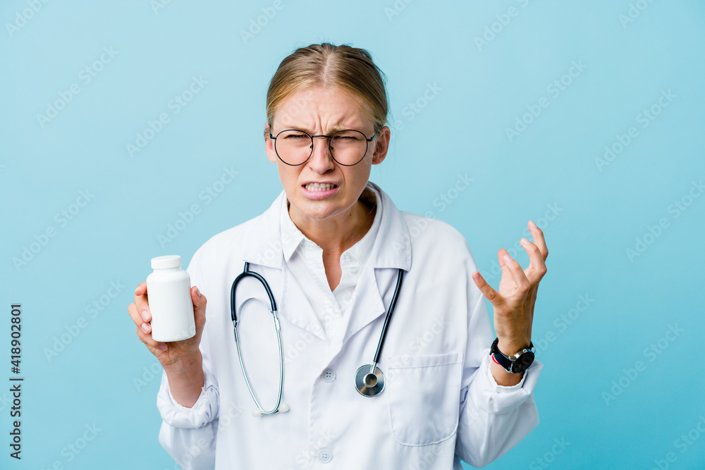 Young russian doctor woman holding pills bottle on blue screaming with rage. Young russian doctor woman holding pills bottle on blue screaming with rage.