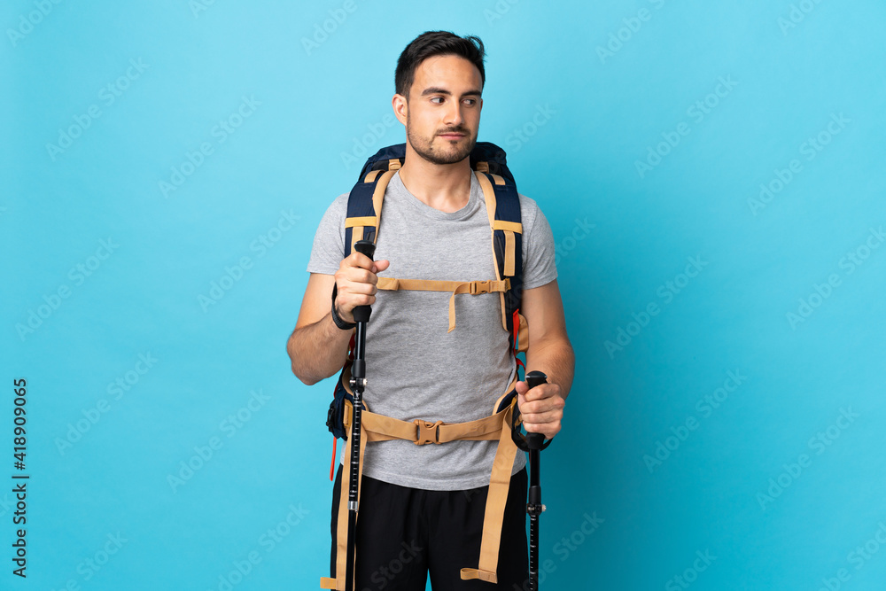 Young caucasian man with backpack and trekking poles isolated on blue background having doubts while looking side