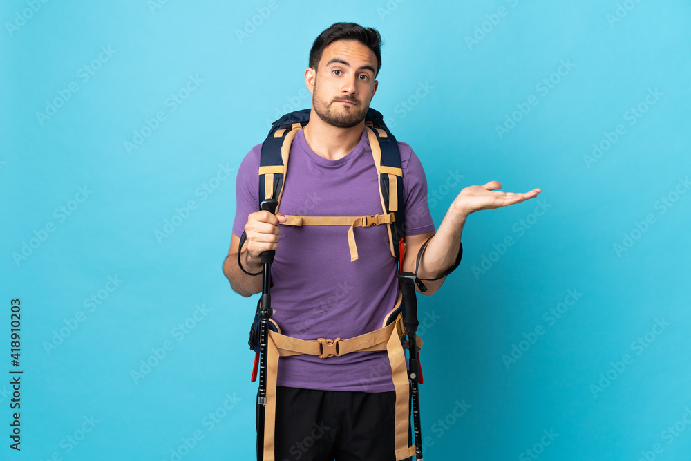 Young caucasian man with backpack and trekking poles isolated on blue background having doubts Young caucasian man with backpack and trekking poles isolated on blue background having doubts