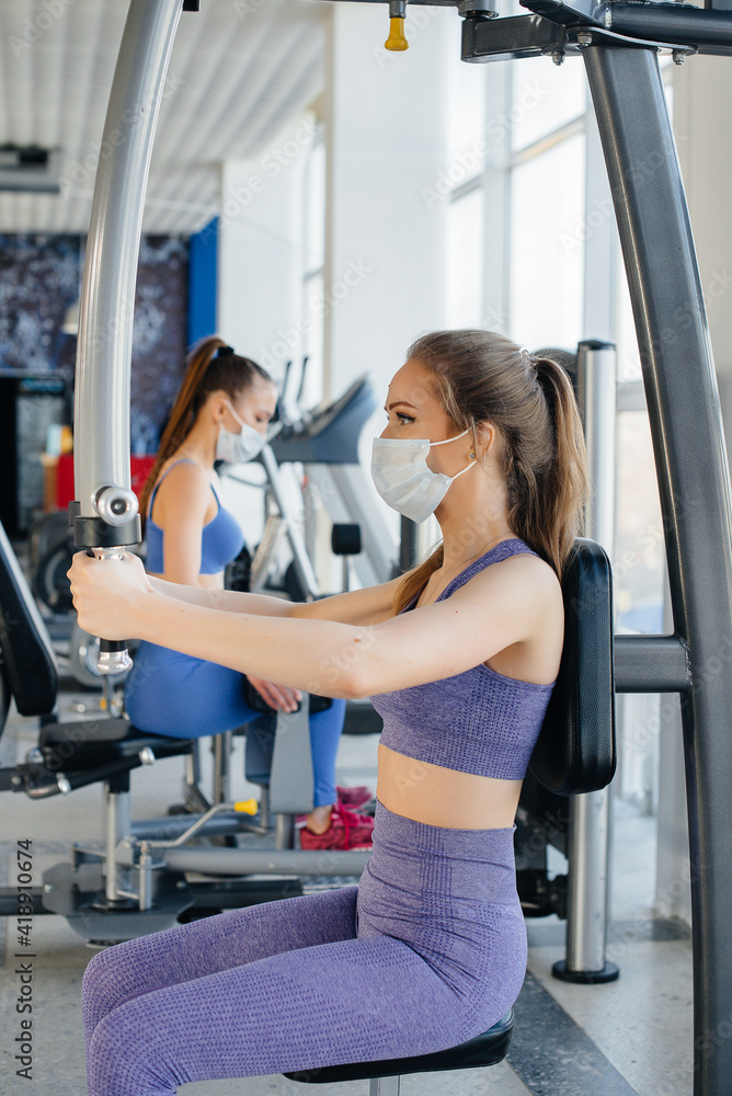 Fototapeta premium Two young beautiful girls exercise in the gym wearing masks during the pandemic. Social distancing in public places.