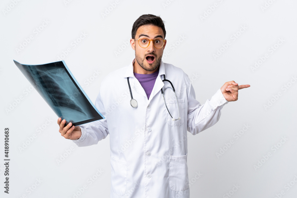 Young doctor man holding a radiography isolated on white background surprised and pointing finger to the side Young doctor man holding a radiography isolated on white background surprised and pointing finger to the side