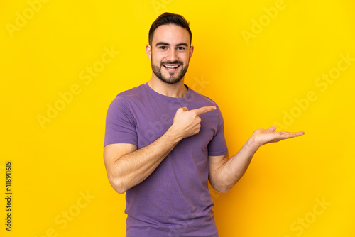 Young caucasian handsome man isolated on yellow background holding copyspace imaginary on the palm to insert an ad