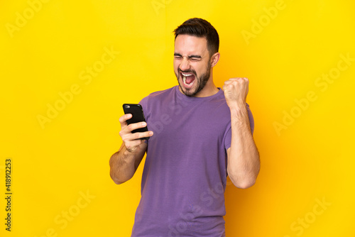 Young caucasian handsome man isolated on yellow background with phone in victory position