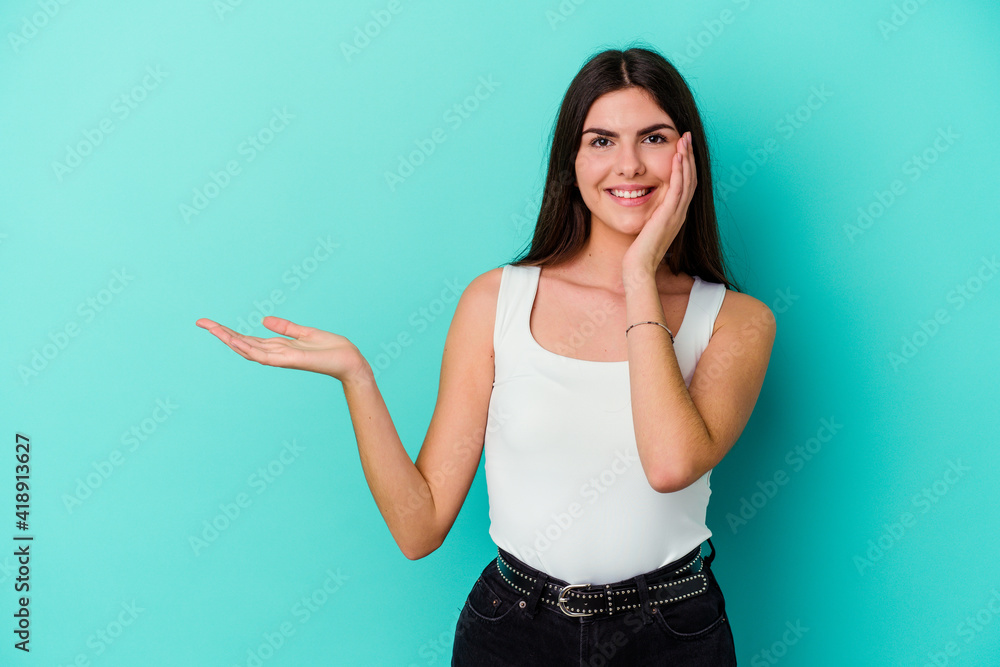 Young caucasian woman isolated on blue background holds copy space on a palm, keep hand over cheek. Amazed and delighted. Young caucasian woman isolated on blue background holds copy space on a palm, keep hand over cheek. Amazed and delighted.