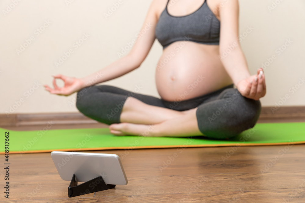 Pregnant female holding smartphone while sitting on exercise yoga mat at home. Fitness or yoga training online at coronavirus time