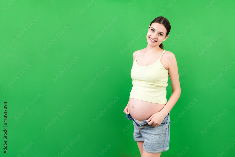 Happy pregnant woman in unzipped jeans showing her naked abdomen at colorful background with copy space Happy pregnant woman in unzipped jeans showing her naked abdomen at colorful background with copy space