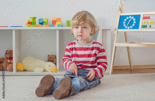 Cute little child using a smartphone while sitting on the floor at home. Digital device and screen time addiction, technology in the hands of children concept.